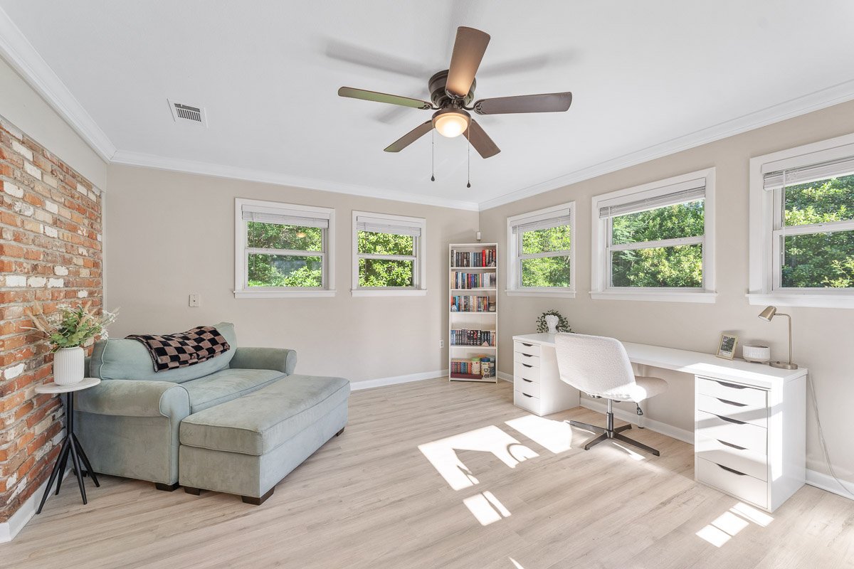 Real Estate photo of a bright home office with four windows, a white desk with drawers, a white office chair, a bookcase, a light green sofa, a small side table with a plant, and a ceiling fan.
