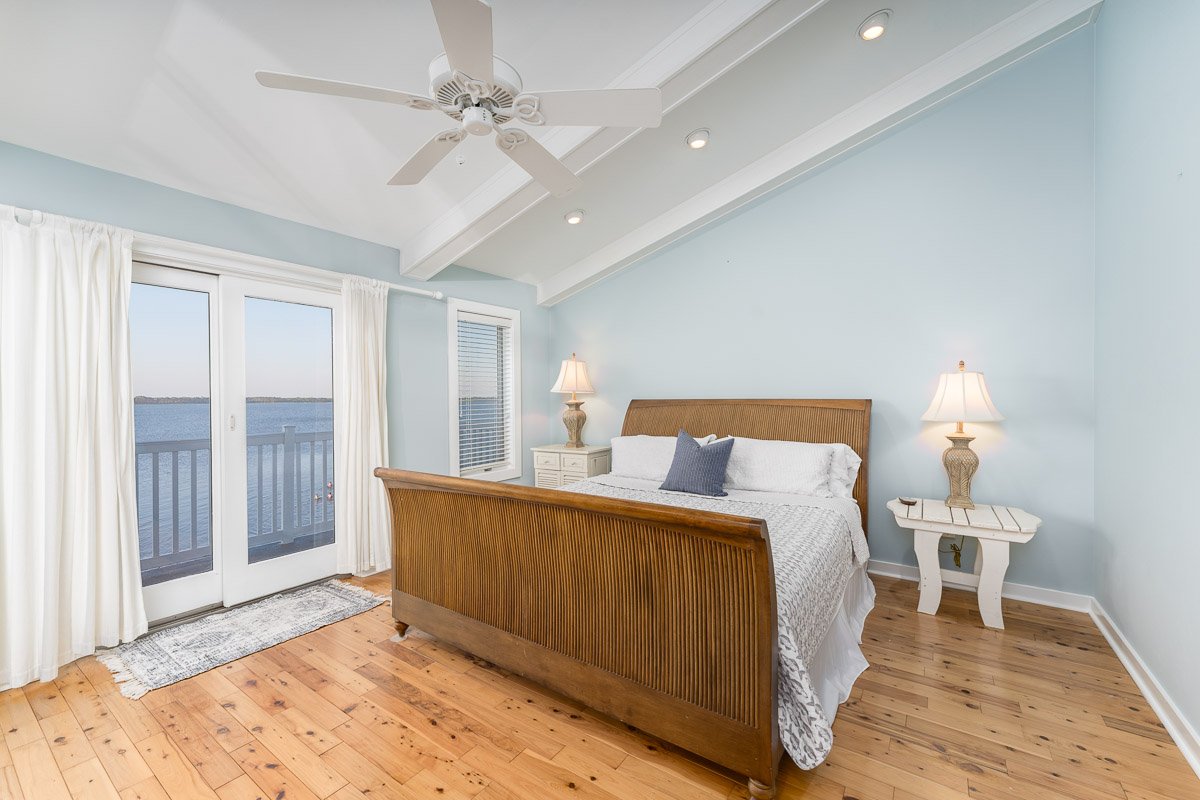 Real Estate photo of a beachhouse bedroom with light blue walls, wooden bed with white bedding and a gray pillow, two white nightstands with lamps, sliding glass door with white curtains leading to a balcony with water view, and hardwood floors.