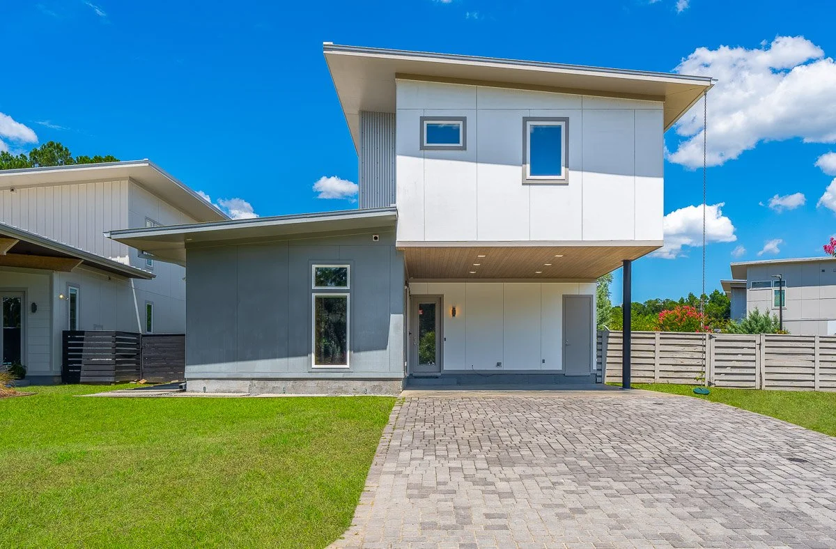 Real Estate photo of a modern two-story house with a gray and white exterior, large windows, and a driveway made of stone pavers, under a blue sky with clouds.