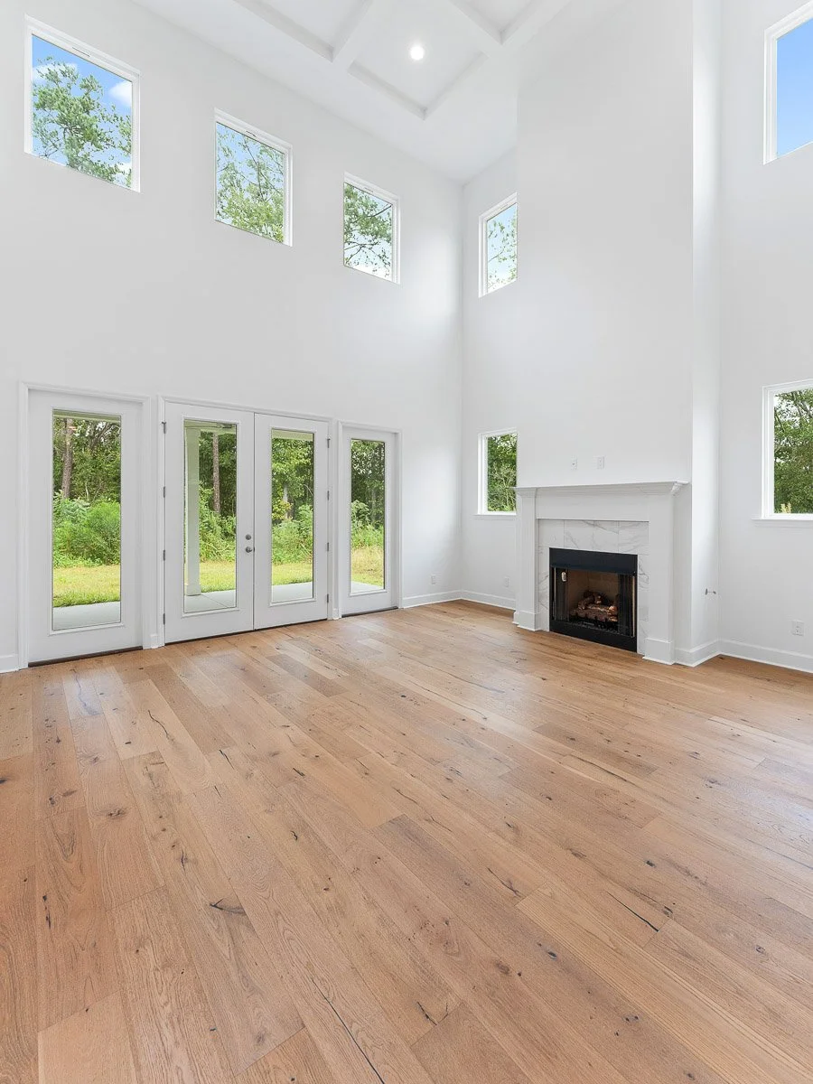 Real Estate photo of a living room with white walls, 20ft ceiling, a fireplace, large windows, and hardwood floors, overlooking a green outdoor area.