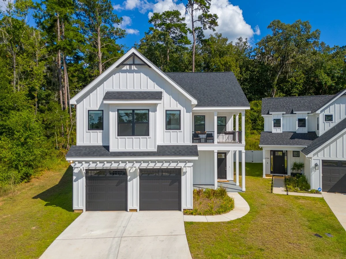 Low aerial Real Estate photo of a newly-built two-story house with white exterior siding, black garage doors, and a black roof, situated in a suburban neighborhood with green trees and a blue sky.
