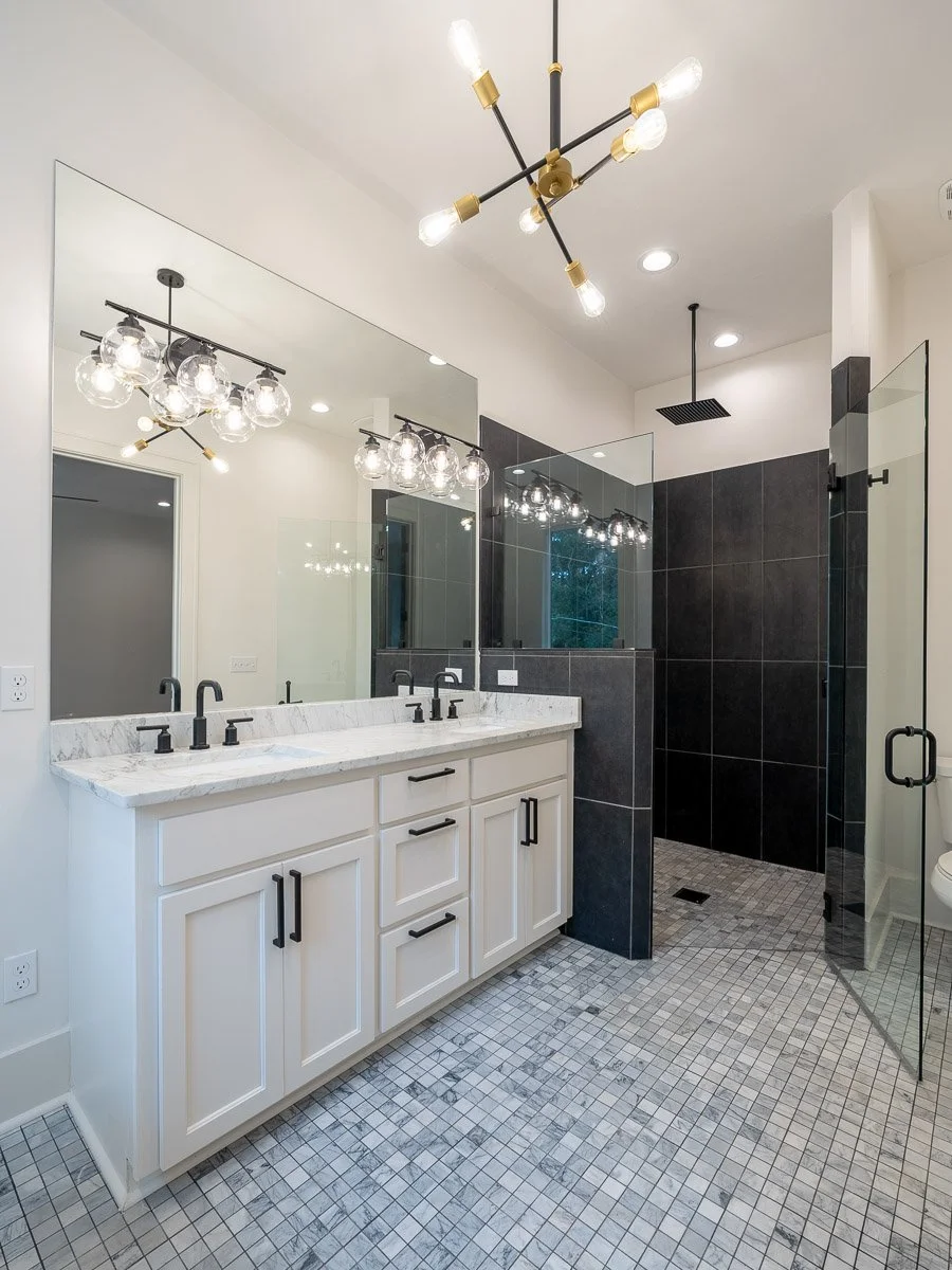 Real Estate photo of modern bathroom with white double vanity, black hardware, large mirror, black tiled shower with glass enclosure, and patterned floor tiles lighting by industrial-style ceiling lights.