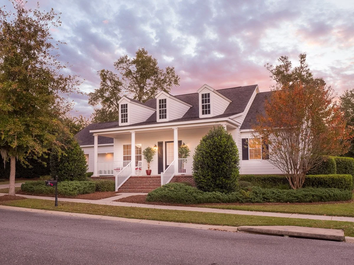 Real Estate photo of a white home with a covered front porch, black front door, front steps, surrounded by trees and bushes, under a colorful cloudy sky during sunset.