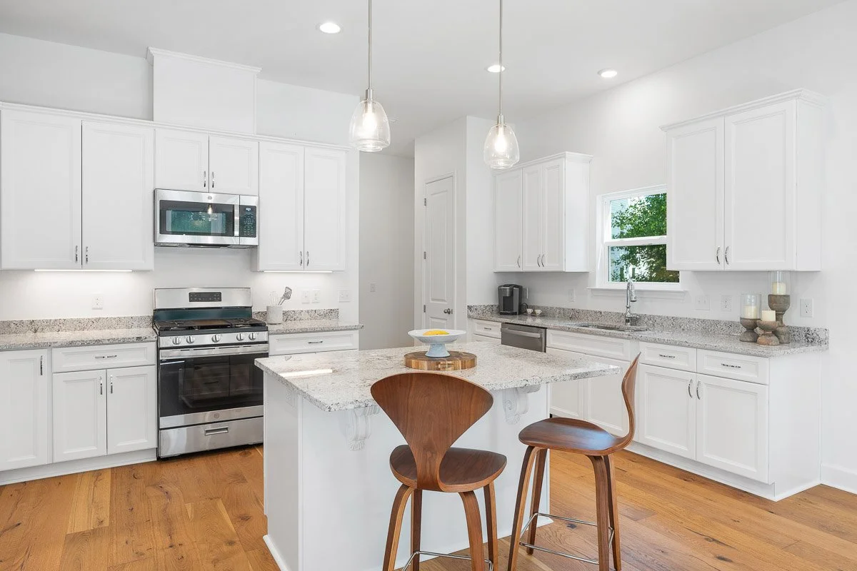 Real Estate photo of a modern white kitchen with granite countertops, stainless steel appliances, and wooden bar stools.