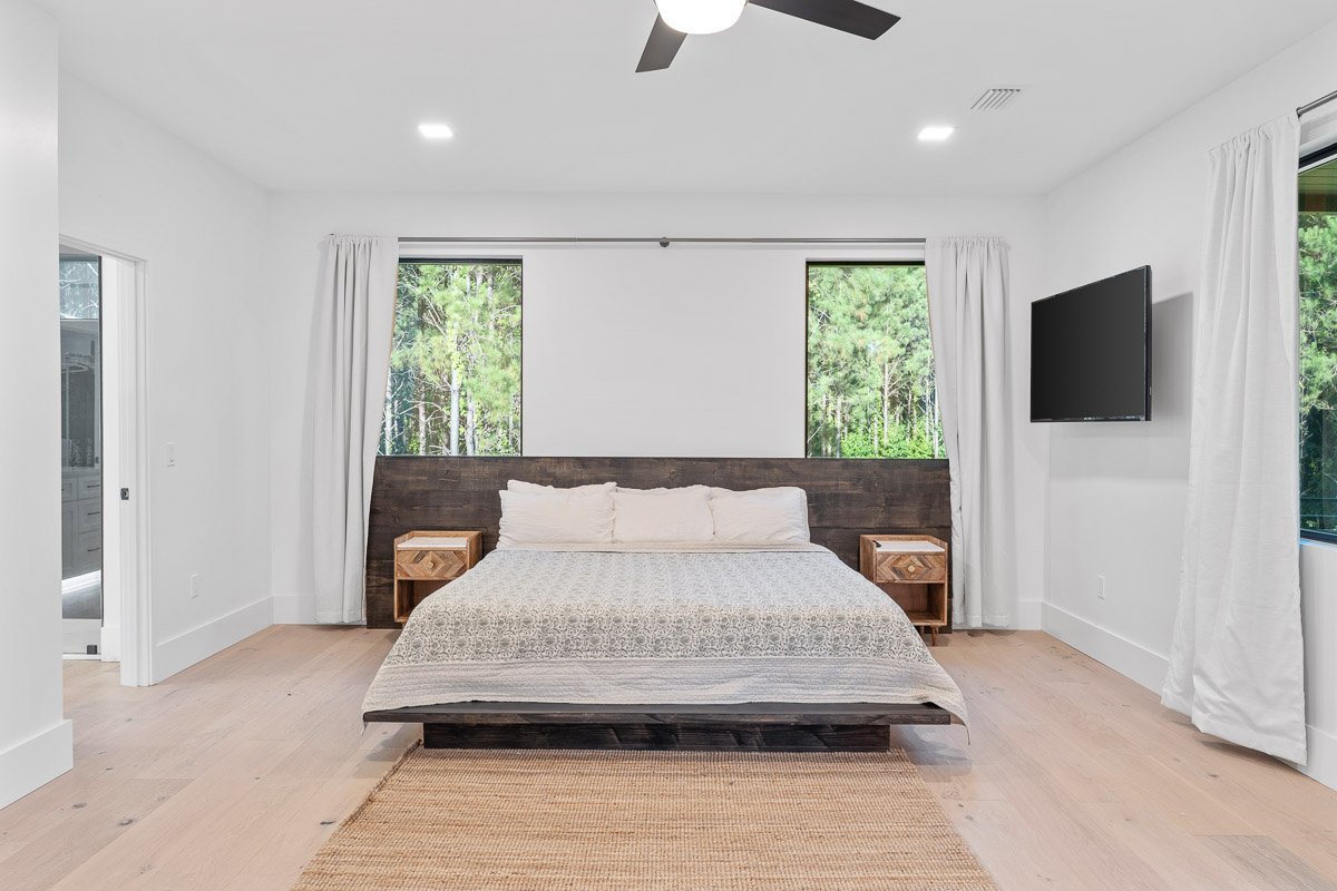 Real Estate photo of a minimalist bedroom with a bed, two nightstands, and a wall-mounted flat-screen TV, featuring large windows with white curtains overlooking trees.