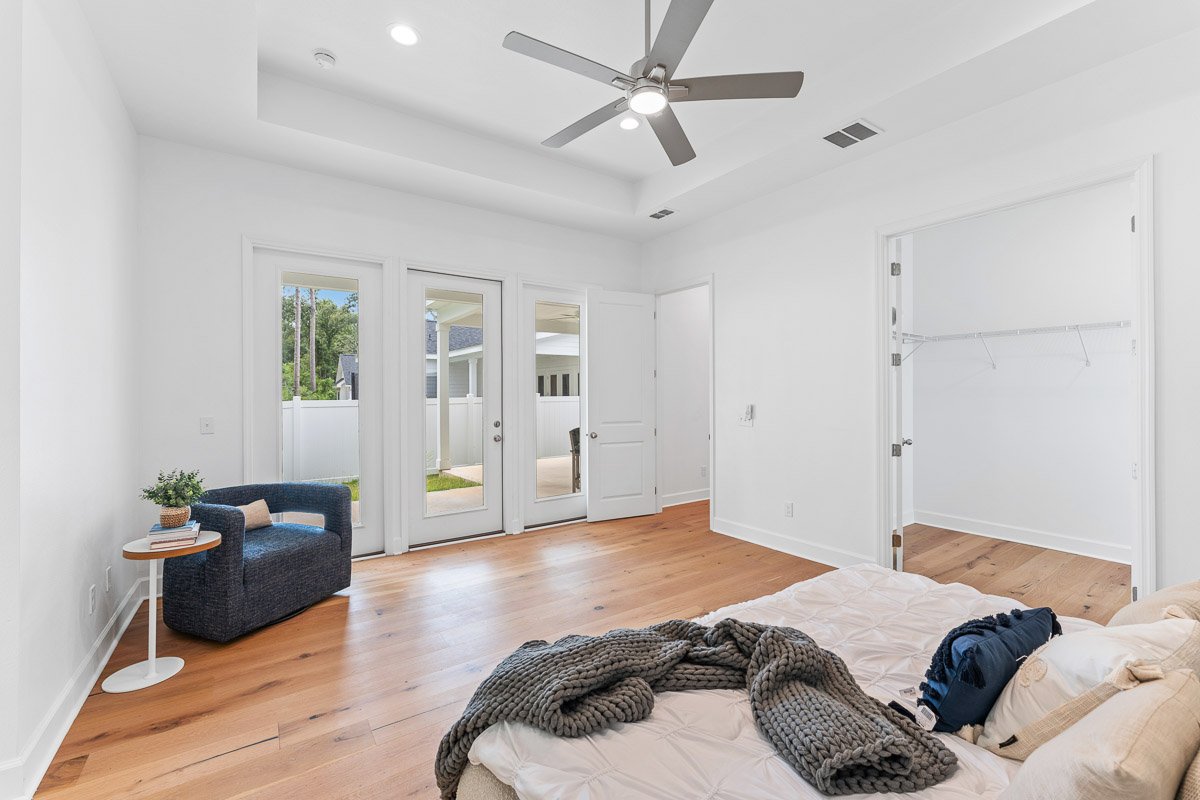 Real Estate photo of a minimally staged bedroom with white walls, hardwood floor, ceiling fan, and glass doors leading outside. There is a small blue chair with a side table and a blanket on the bed.