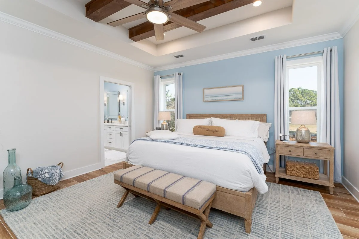 Bright bedroom with a large bed, white bedding, and a light blue accent wall. There are two windows with white curtains, a wooden nightstand with a lamp and decorative vase, a bench at the foot of the bed, and an open bathroom door revealing a vanity
