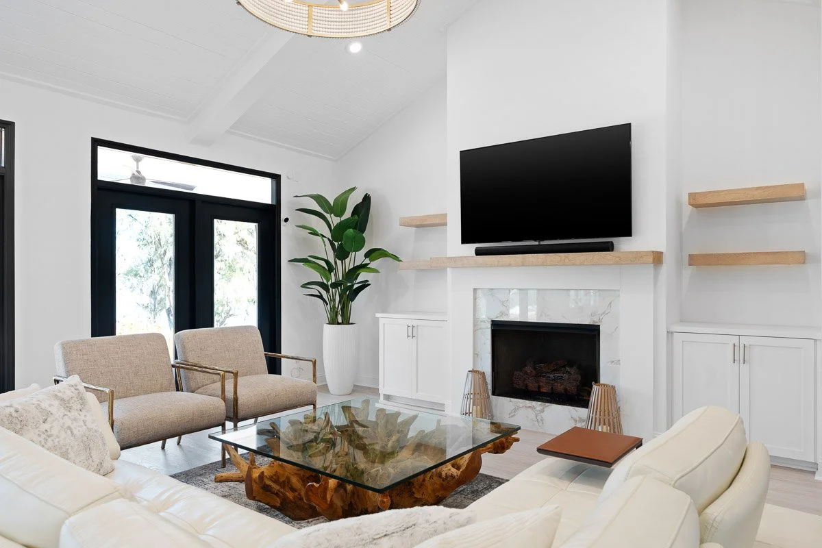 Real Estate photo of a modern living room with white walls, a fireplace, a wall-mounted TV, beige and white seating, a glass coffee table with a natural wood base, and large windows with black frames.