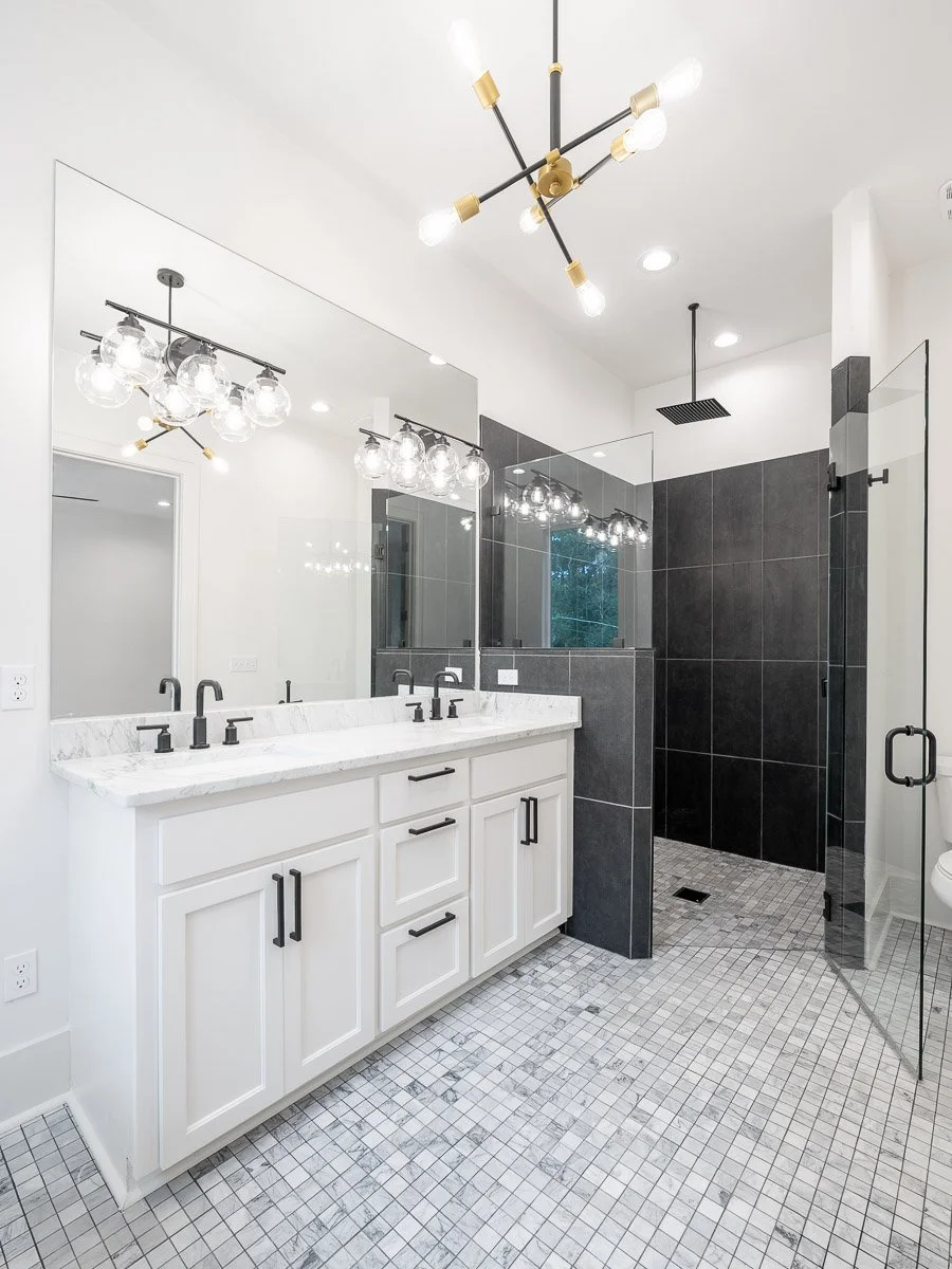 Real Estate photo of modern bathroom with white double vanity, black hardware, large mirror, black tiled shower with glass enclosure, and patterned floor tiles lighting by industrial-style ceiling lights.