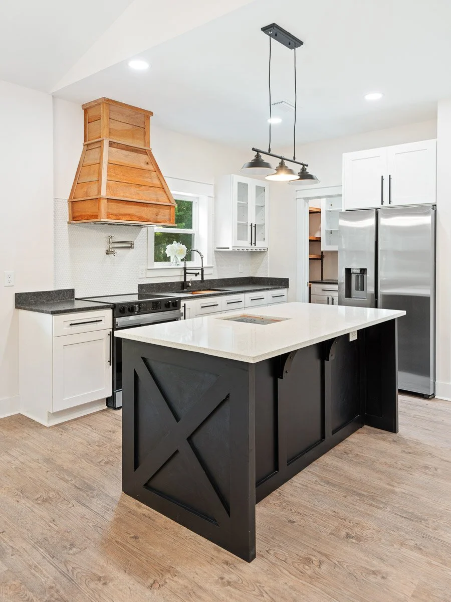 Real Estate photo of a modern kitchen with black island, white cabinets, stainless steel refrigerator, black countertops, wooden range hood, and pendant lighting.