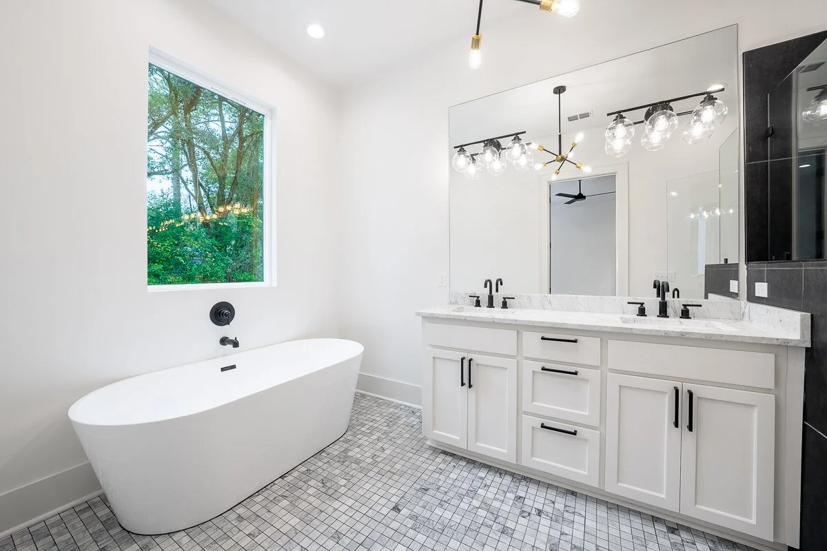 Real Estate photo of a modern bathroom with white stand-alone bathtub, large mirror, double sink vanity, black fixtures, and a window showing greenery outside.