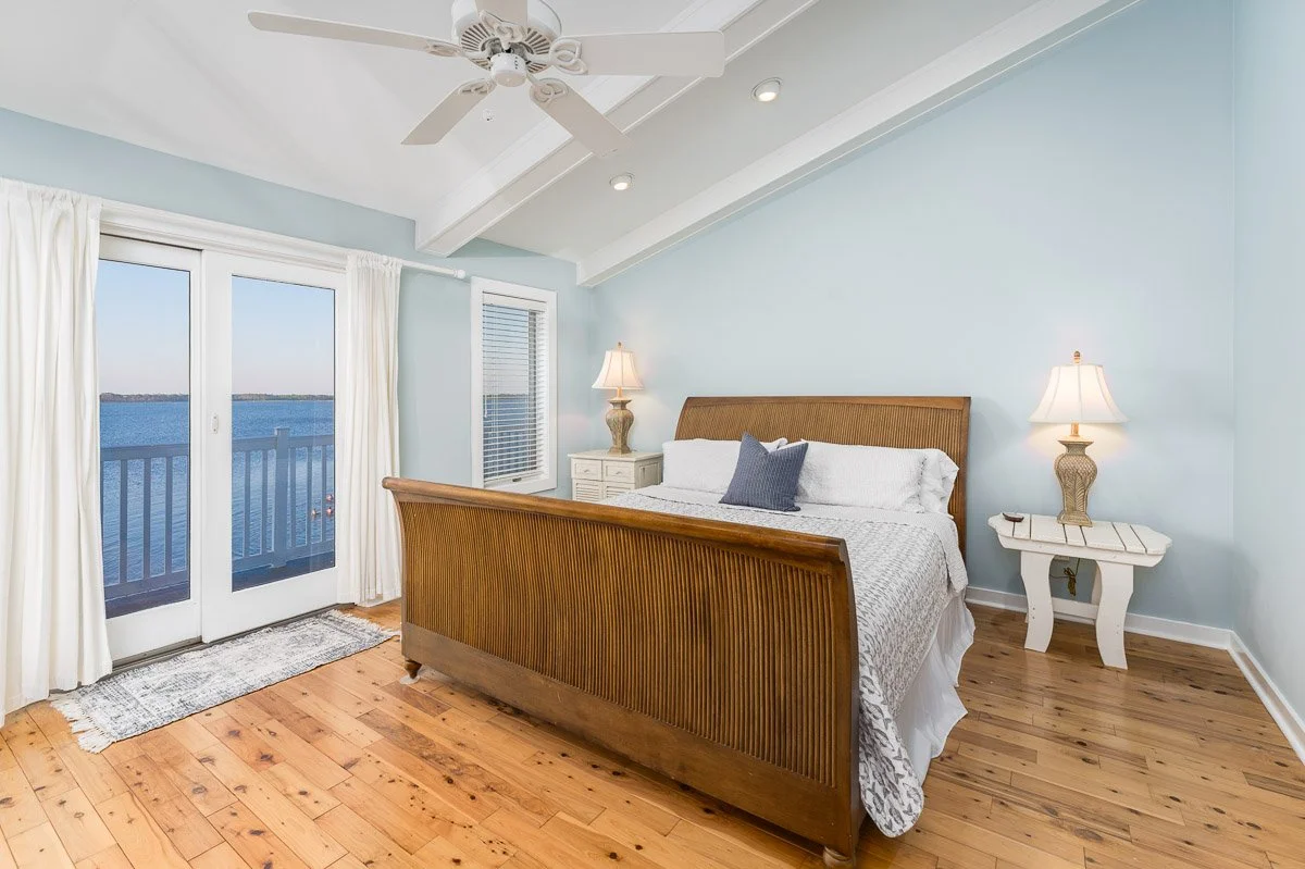 Real Estate photo of a beachhouse bedroom with light blue walls, wooden bed with white bedding and a gray pillow, two white nightstands with lamps, sliding glass door with white curtains leading to a balcony with water view, and hardwood floors.