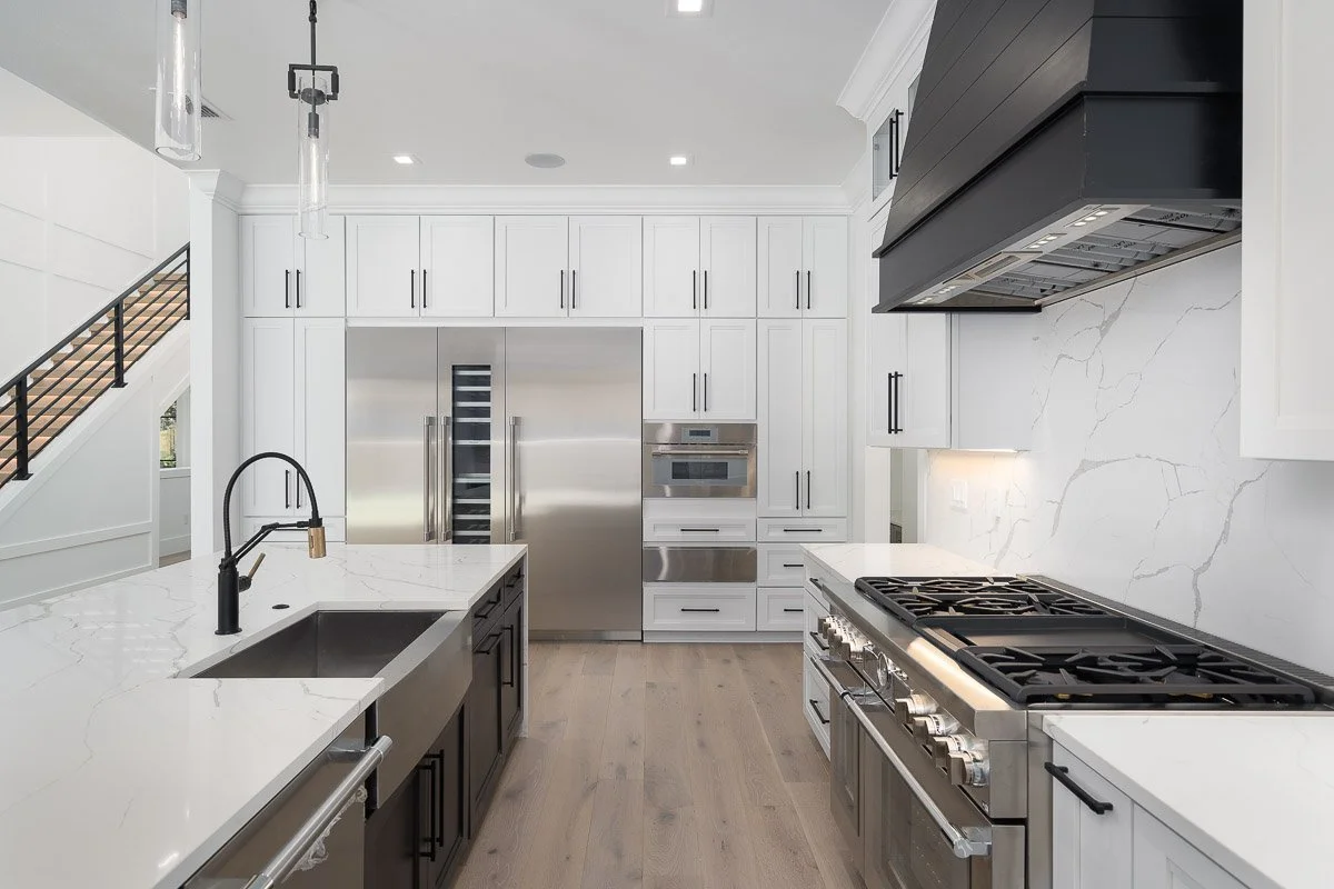 Modern kitchen with white cabinetry, stainless steel appliances, a black range hood, a marble backsplash, and a kitchen island with a black faucet.