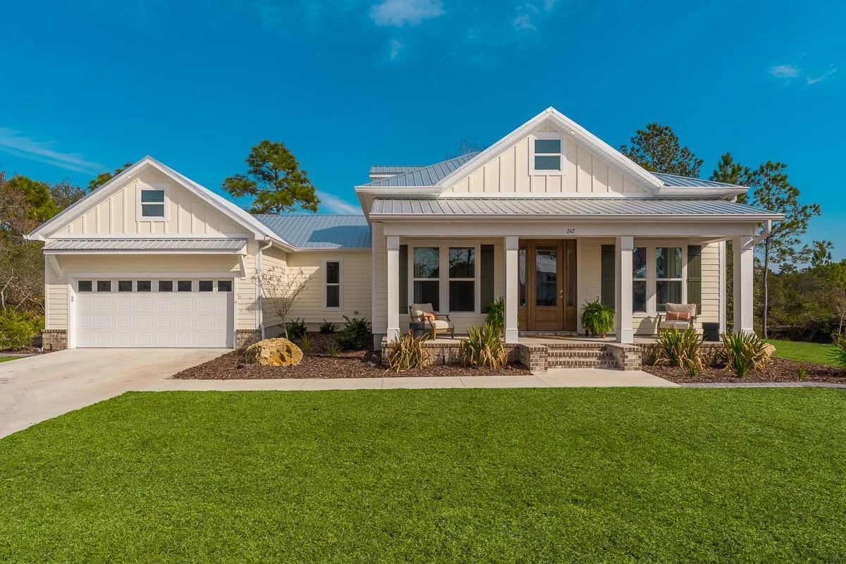 Front view of a two-story house with a white exterior, gray metal roof, front porch with plants, and a attached garage on a landscaped lawn under a blue sky.
