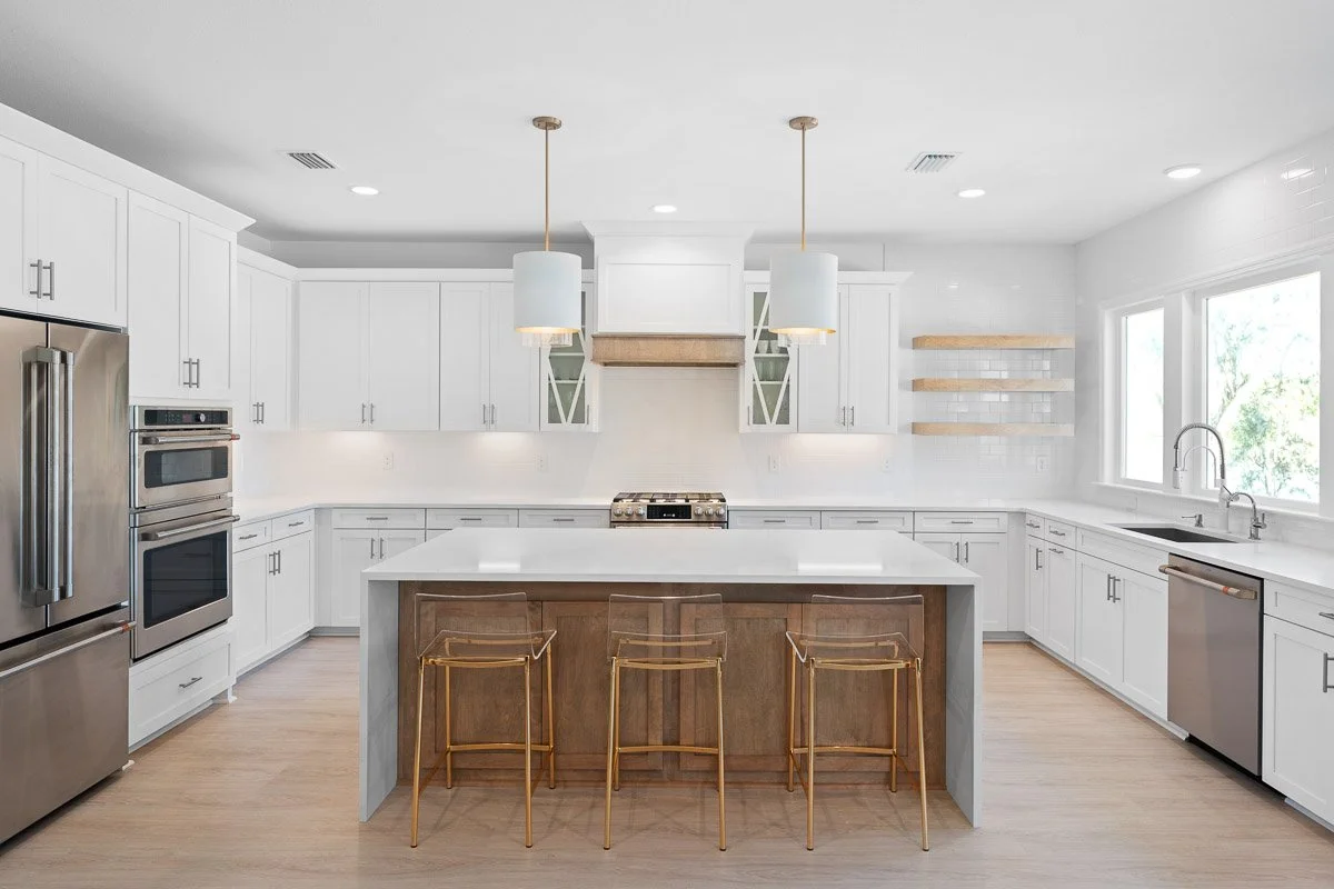 Real Estate photo of a bright white kitchen with an island, wooden accents, gold bar stools, stainless steel appliances, open shelving, and large windows.