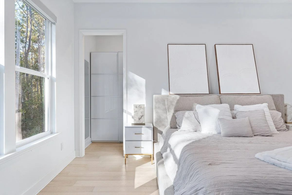 Architectural style Real Estate photo of a bright and airy bedroom with large window, white walls, beige upholstered bed with multiple pillows, and a small white nightstand with gold legs and a marble lamp.
