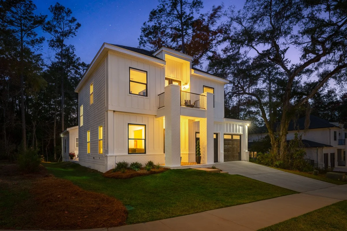 Real Estate photo of a modern two-story white house at dusk with illuminated windows, a front porch, and a driveway leading to a garage, surrounded by trees and a well-maintained lawn.