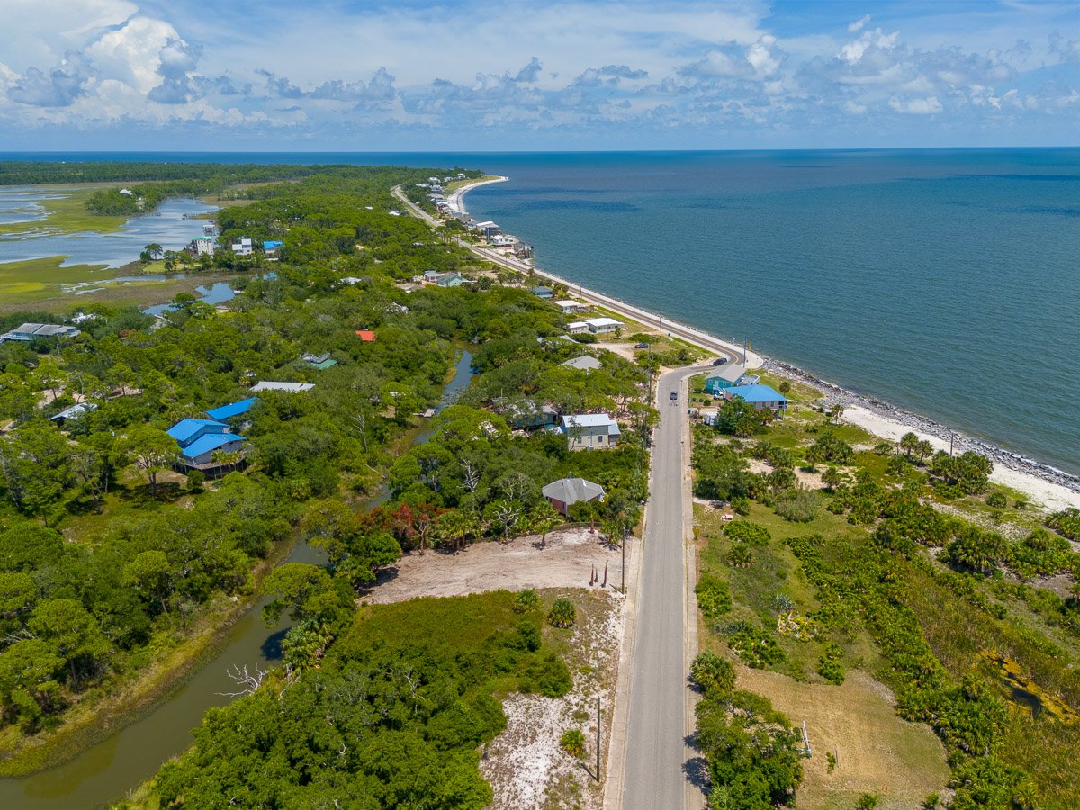 Aerial Real Estate photo of a cleared, empty lot of land on Alligator Point, surrounded by green vegetation, a waterway, and the ocean with a partly cloudy sky.