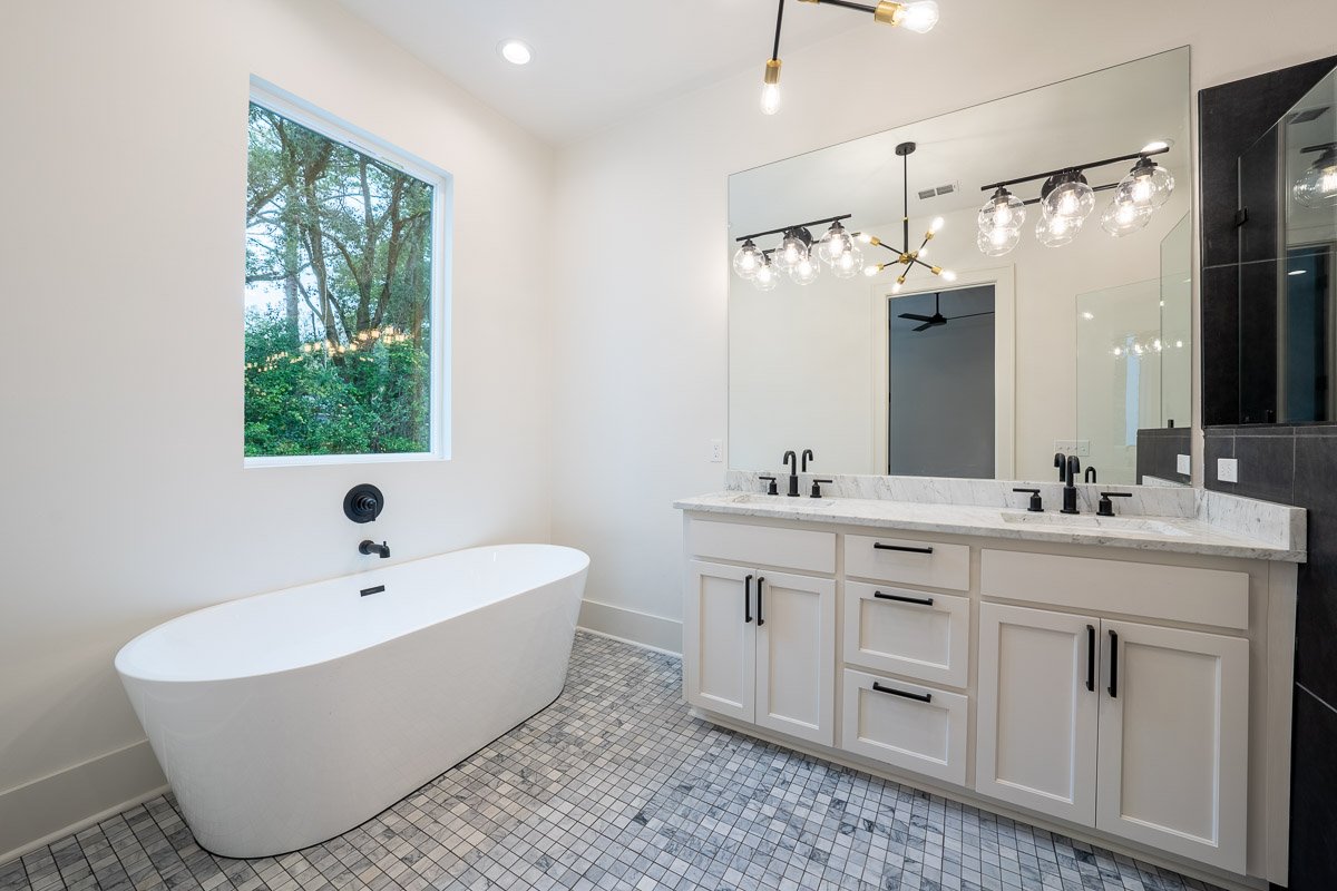 Real Estate photo of a modern bathroom with white stand-alone bathtub, large mirror, double sink vanity, black fixtures, and a window showing greenery outside.