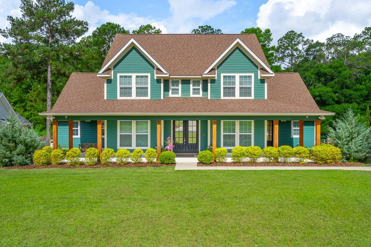 Real Estate photo of a a two-story house with a green exterior, brown roof, white window shutters, and a front yard with neatly trimmed bushes and green lawn.