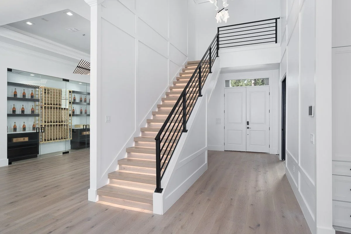 Interior of a modern home entryway with a staircase featuring black metal railing, white walls, light wood flooring, and a front door. There is a room with glass doors to the left and a small window above the door.