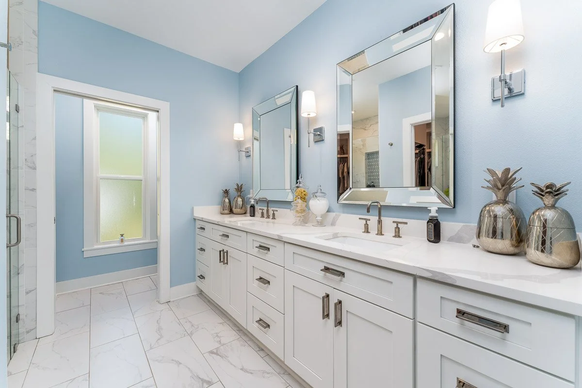Bathroom with blue walls, white vanity with two sinks, large framed mirrors, decorative pineapple vases, and modern fixtures.