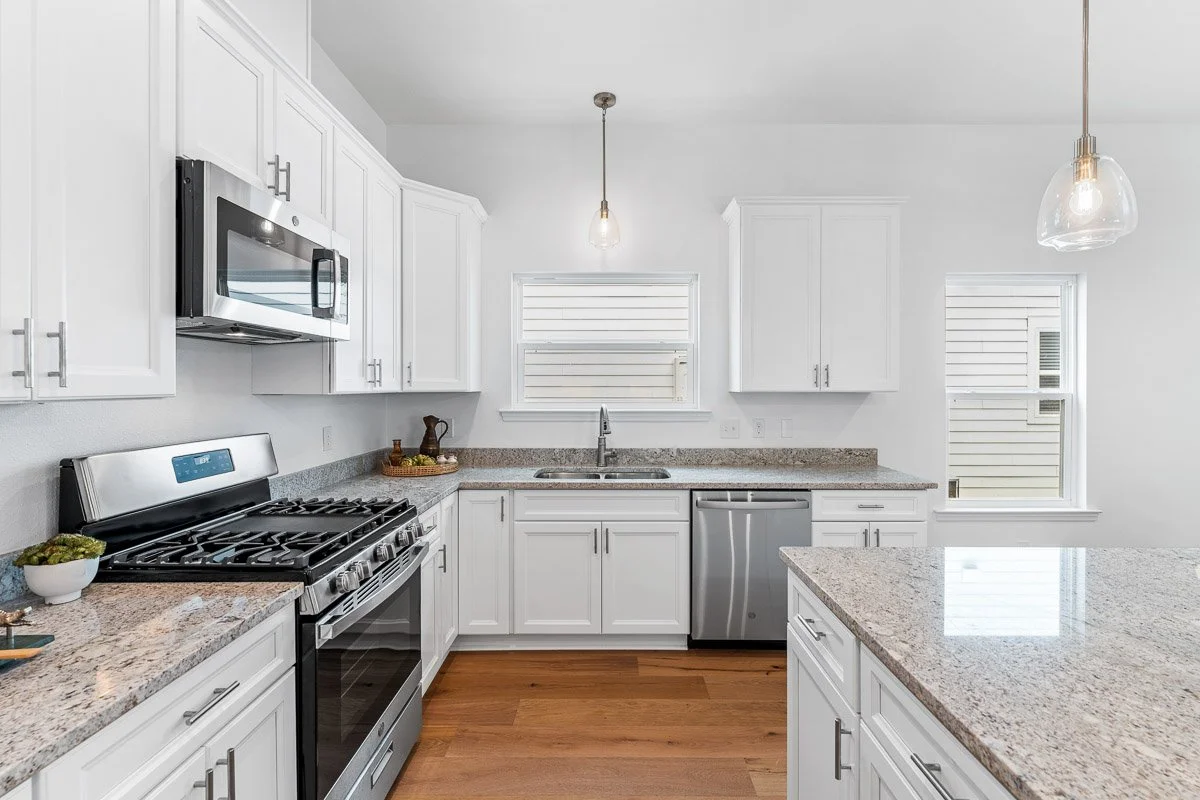 Real Estate photo of a clean white kitchen with granite countertops, stainless steel appliances, hardwood floors, and two windows with blinds.
