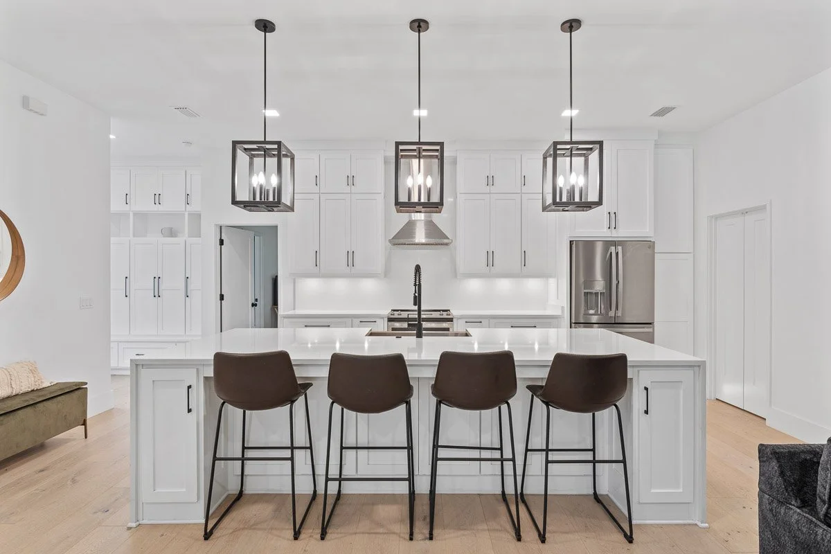 Real Estate photo of a modern white kitchen with a large island, four black barstools, three pendant lights, and stainless steel appliances