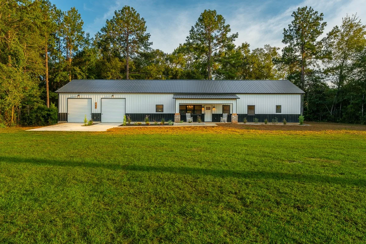 Real Estate photo of a large metal barddominium with two garage doors, a small front porch with chairs, and a grassy yard surrounded by trees under a blue sky.