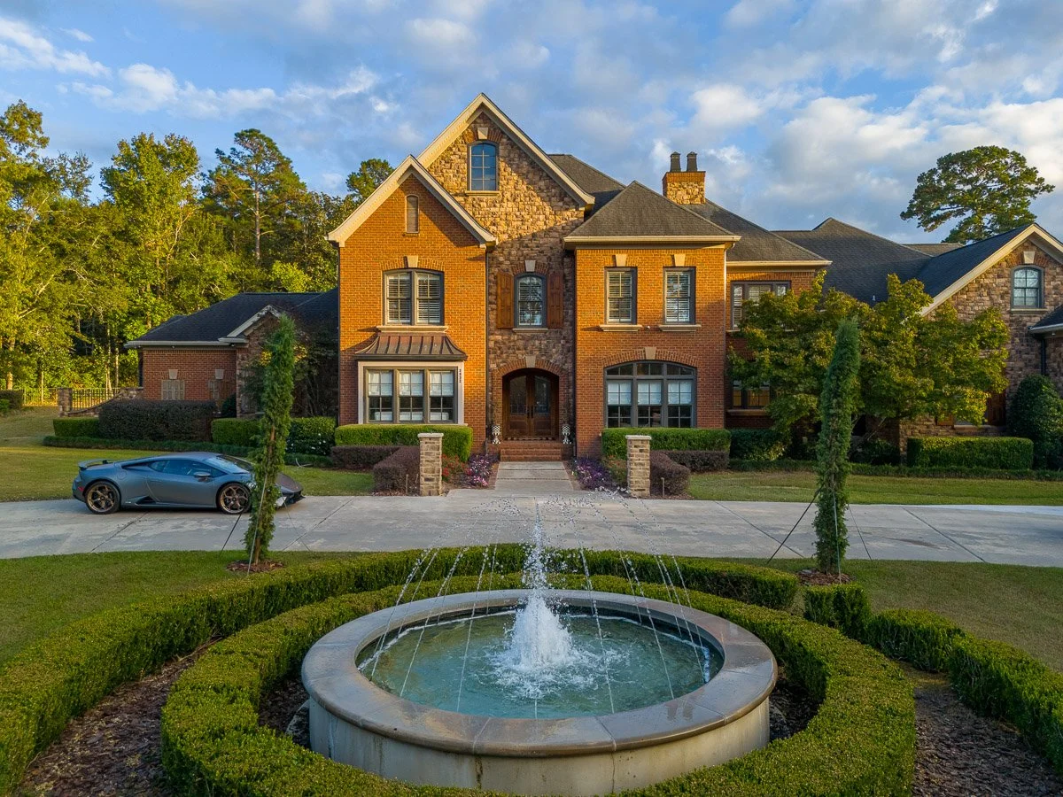 Real Estate photo of a large, elegant brick house with multiple gables and arched windows, a fountain in the foreground, a driveway with a Lamborghini parked on it, and lush green landscaping under a partly cloudy sky.