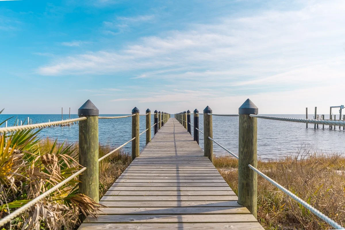 A wooden pier extends over the water with blue sky and clouds above, and grassy dunes on either side.