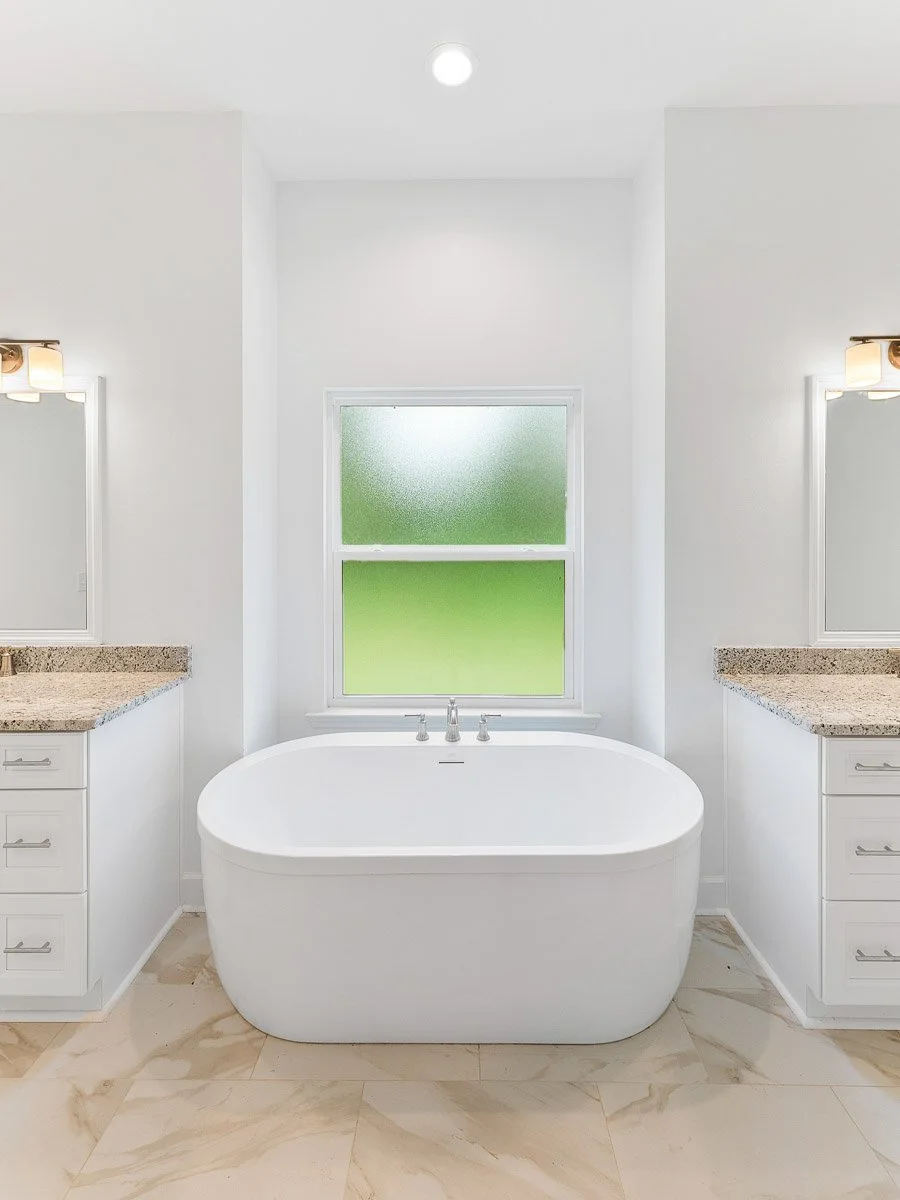 Real Estate photo of a bright bathroom with a freestanding bathtub in front of a frosted window, flanked by two white vanities with granite countertops and surrounded by white walls and marble flooring.
