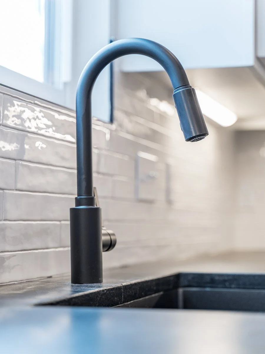 Real Estate detail photo of a  black kitchen faucet over a dark countertop with a tiled backsplash and light-colored upper cabinets in the background.