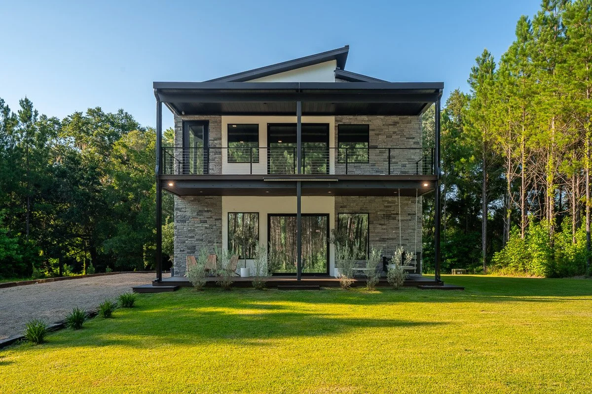 Real Estate photo of a ultra-modern two-story home with stone exterior, large glass doors, and black metal balcony, surrounded by green lawn and trees under a blue sky.