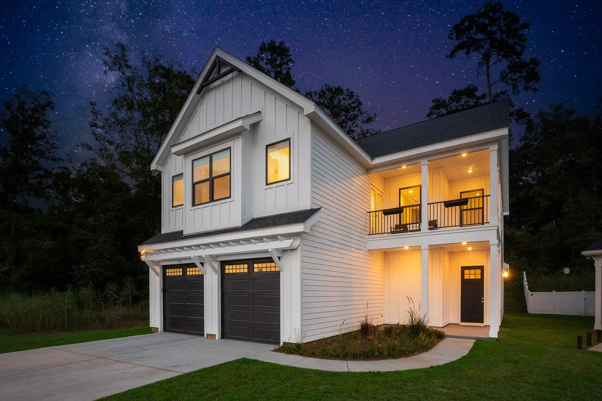 Real Estate photo of a modern white two-story house with black garage doors, lit interior, and a small front porch, under a starry night sky surrounded by trees.