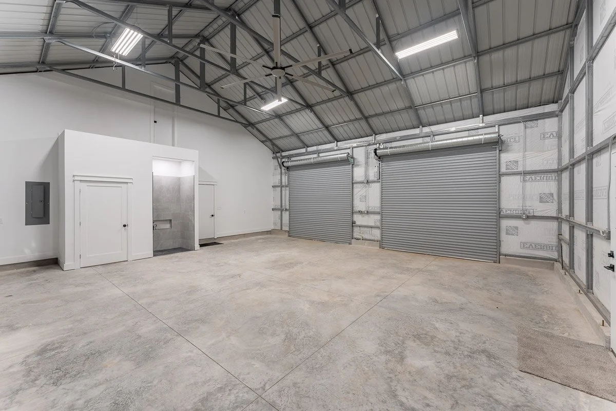Real Estate photo of a barndominium garage with concrete floor, metal walls, high ceiling, and large rolling metal doors.