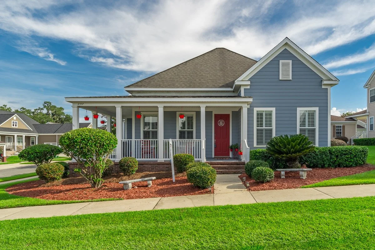 Real Estate photo of the front view of a blue house with a red door, white porch railing, and red hanging flower pots, surrounded by a green lawn and landscaped bushes.