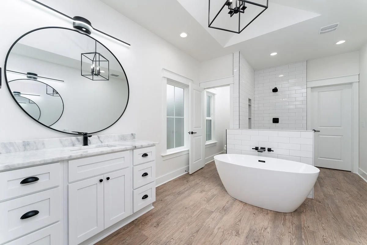 Modern bathroom with white cabinetry, a large round mirror, a freestanding bathtub, white subway tile, and black fixtures.