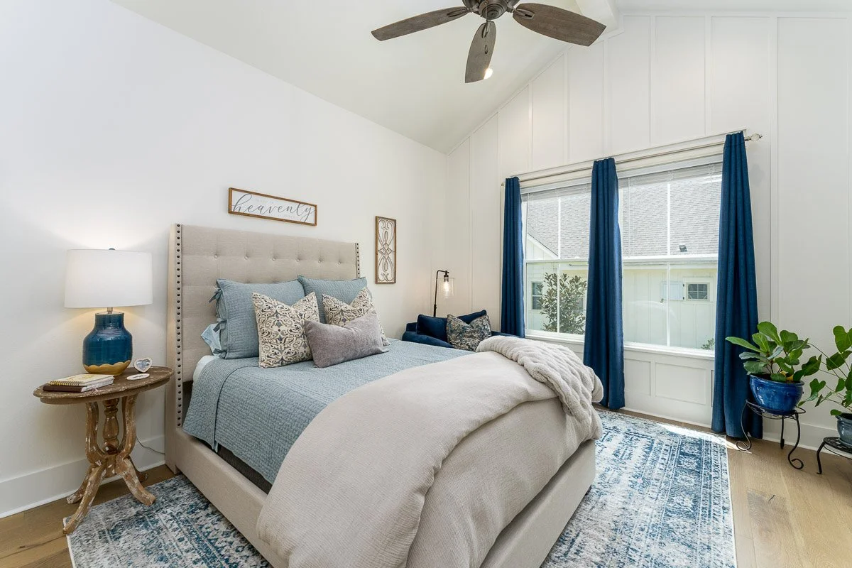 A cozy bedroom featuring a large bed with light-colored upholstered headboard, blue bedding, and decorative pillows. A wooden nightstand with a blue lamp is next to the bed, and there are two windows with blue curtains letting in natural light. There