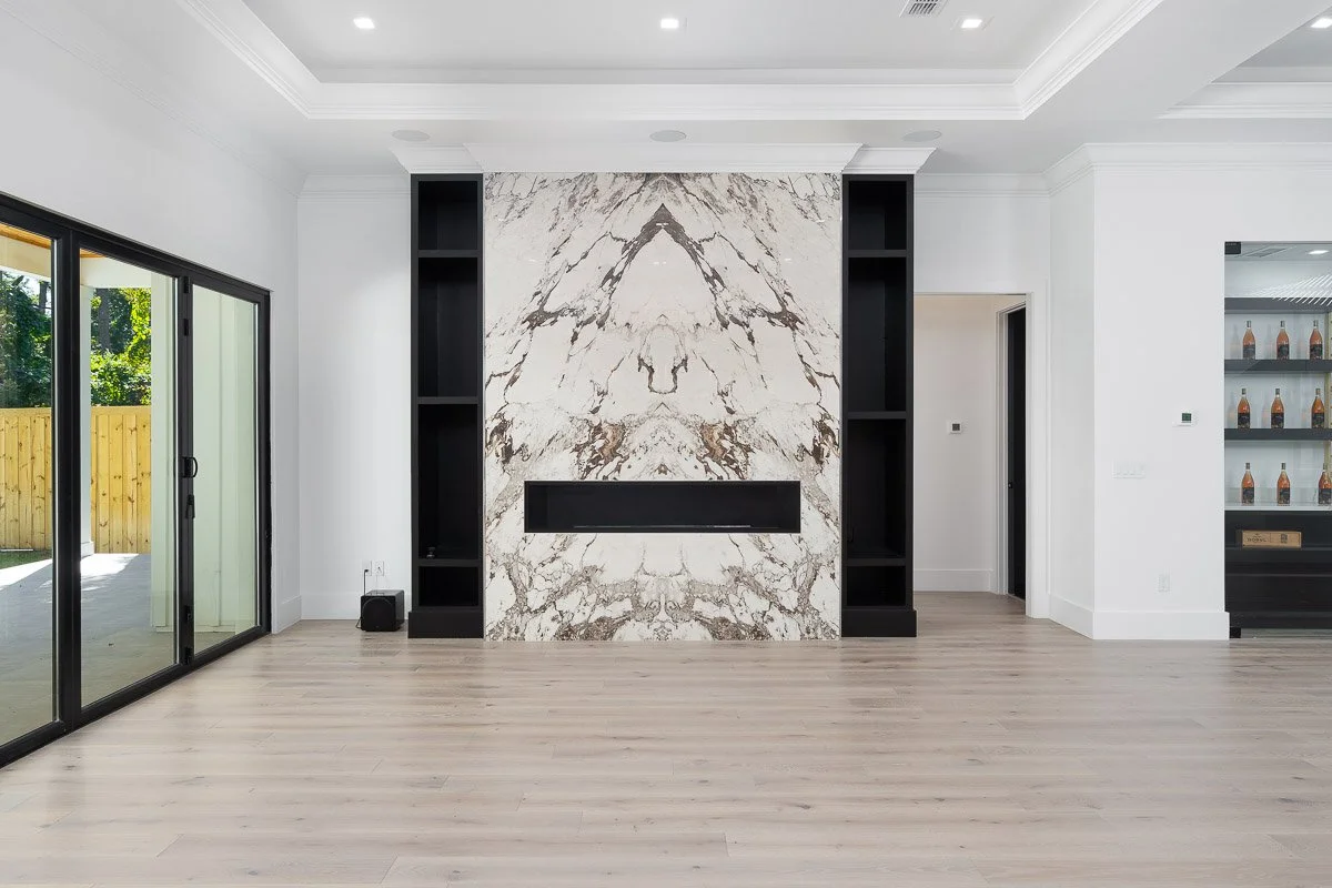 Empty living room with a modern white and black fireplace wall, black shelves on either side, oak flooring, a sliding glass door on the left, and a display cabinet with bottles on the right.