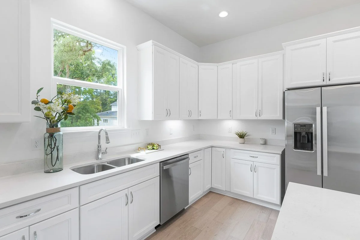 Real Estate photo of a bright white kitchen with large window, white cabinets, stainless steel appliances, light wood flooring, and small decorative plants.
