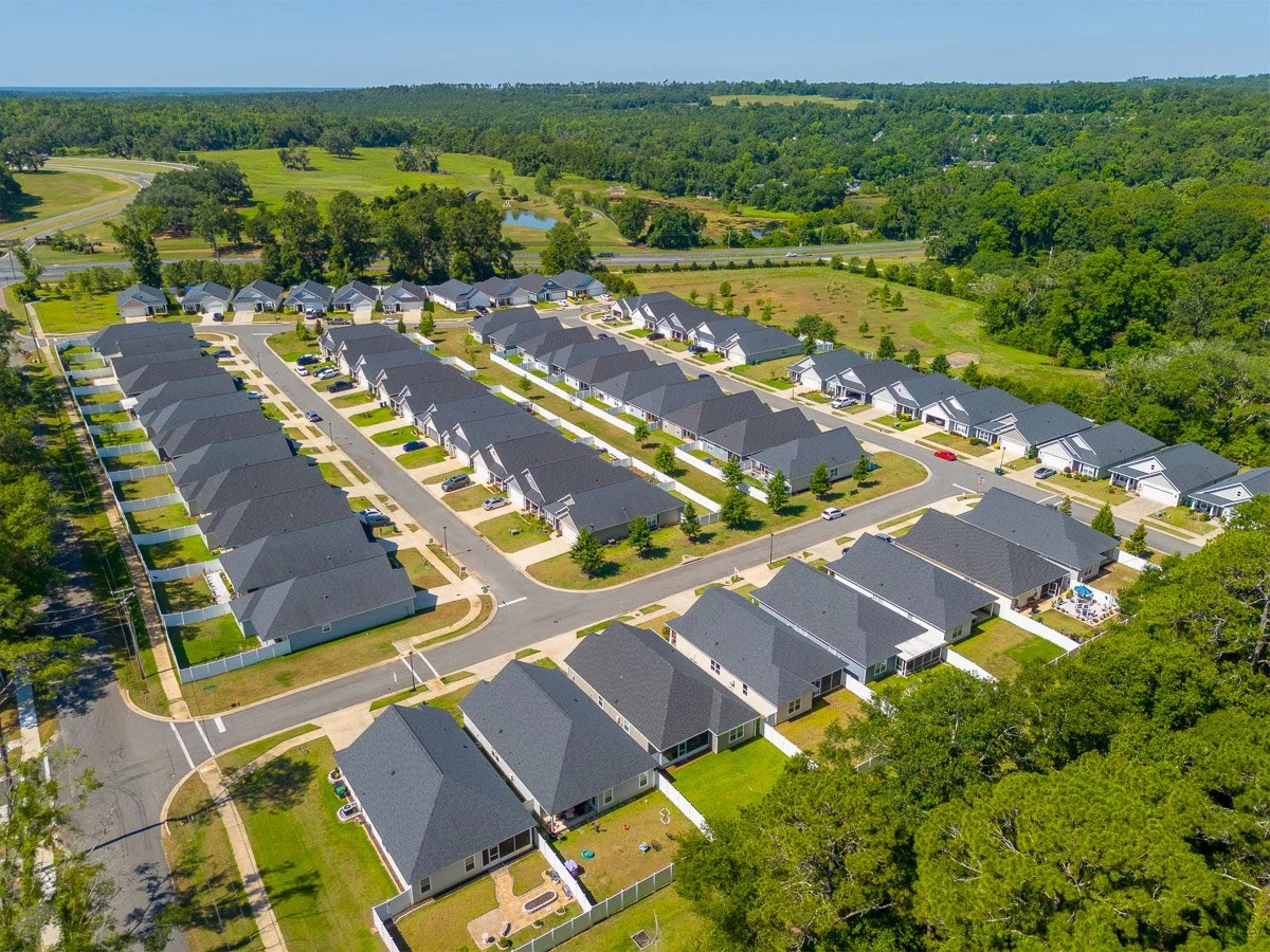 Aerial Real Estate photo of a suburban neighborhood with rows of houses, streets, green lawns, and trees, with a wooded area and a pond in the background.