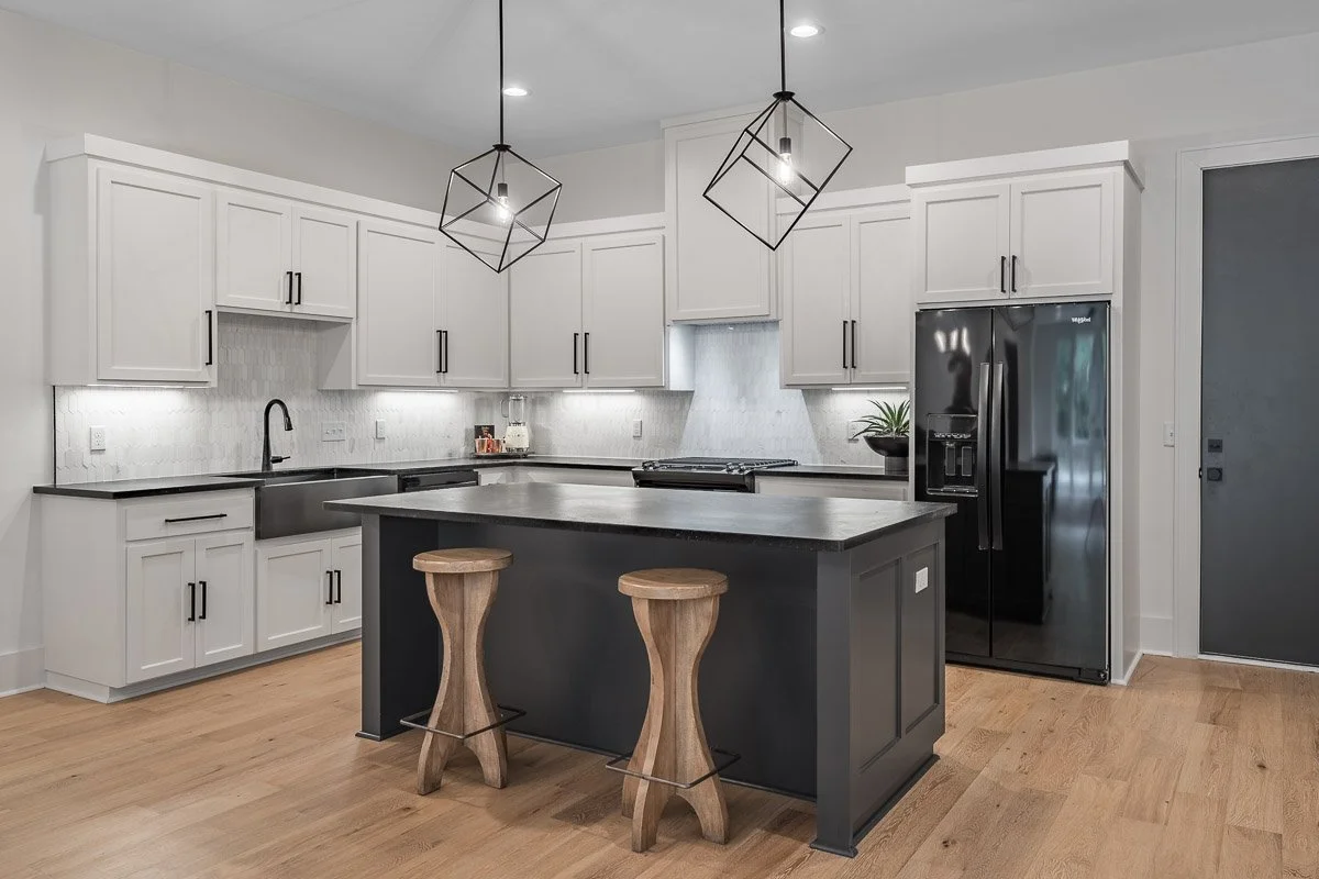 Real Estate photo of a modern kitchen with white cabinets, black countertops, a black refrigerator, and two wooden bar stools at a black kitchen island. There are geometric pendant lights hanging from the ceiling.