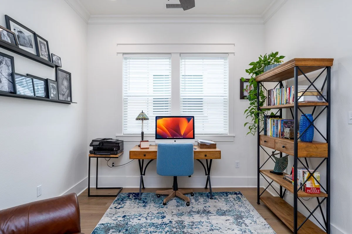 Real Estate photo of a home office with a wooden desk, a blue office chair, a black bookshelf filled with books and decorations, a window with white blinds, a gallery wall of black-and-white photographs.