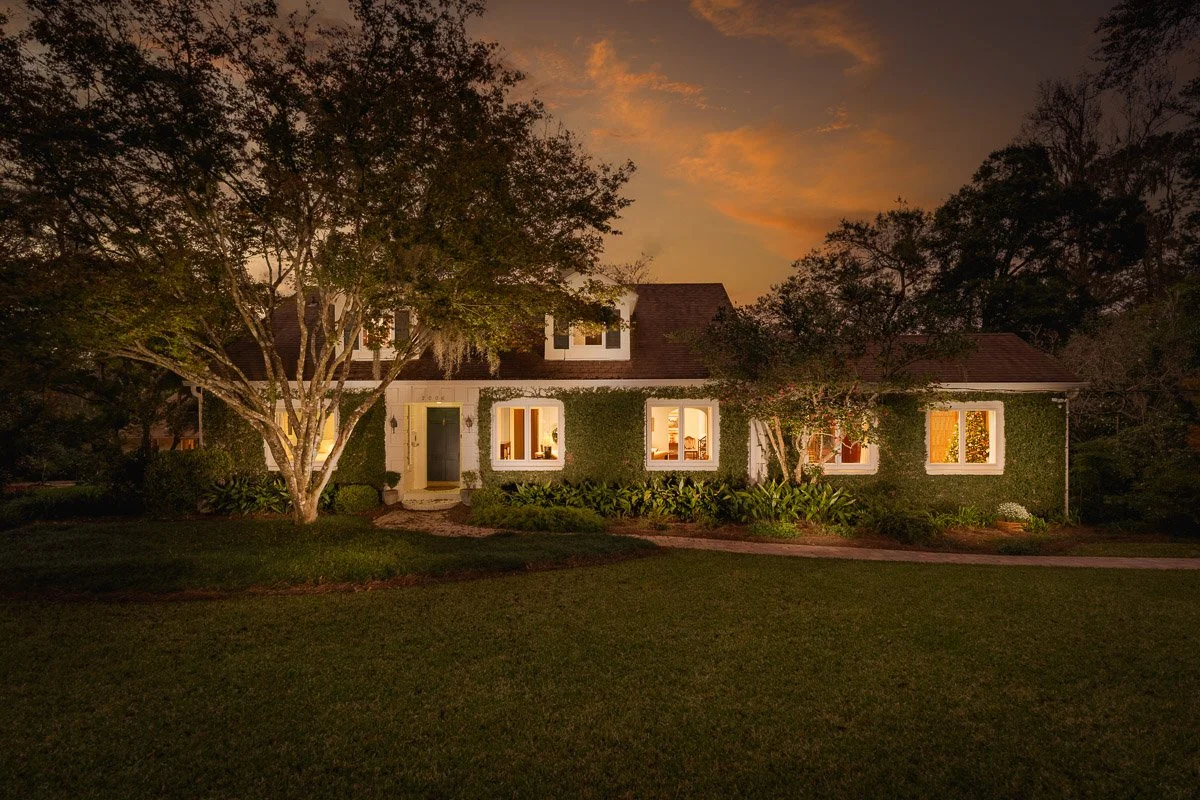Real Estate photo of a mid-century home at sunset with ivy growing on the walls, lights on inside, surrounded by trees and a well-maintained lawn.