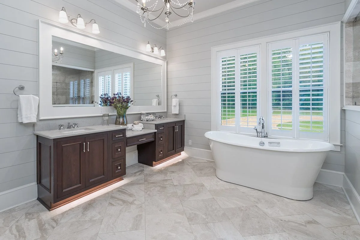 Real Estate photo of a modern bathroom with a white bathtub near a large window with plantation shutters, a dark wood vanity with a large mirror, a chandelier, and neutral floor tiles.