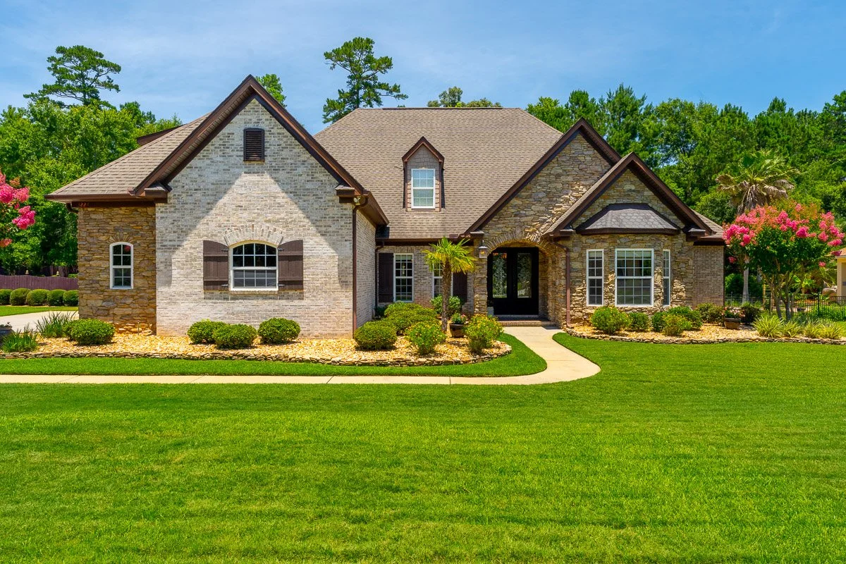 Real Estate photo of a large brick home with a well-maintained front yard, green grass, shrubbery, and trees under a blue sky.