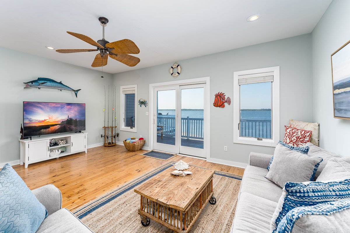 Real Estate photo of a living room in an ocean front condo with waterfront view, featuring a gray sofa with pillows, a wooden coffee table, a TV stand with a large TV, fishing poles, fishing-themed decor, and sliding glass doors leading to a balcony 