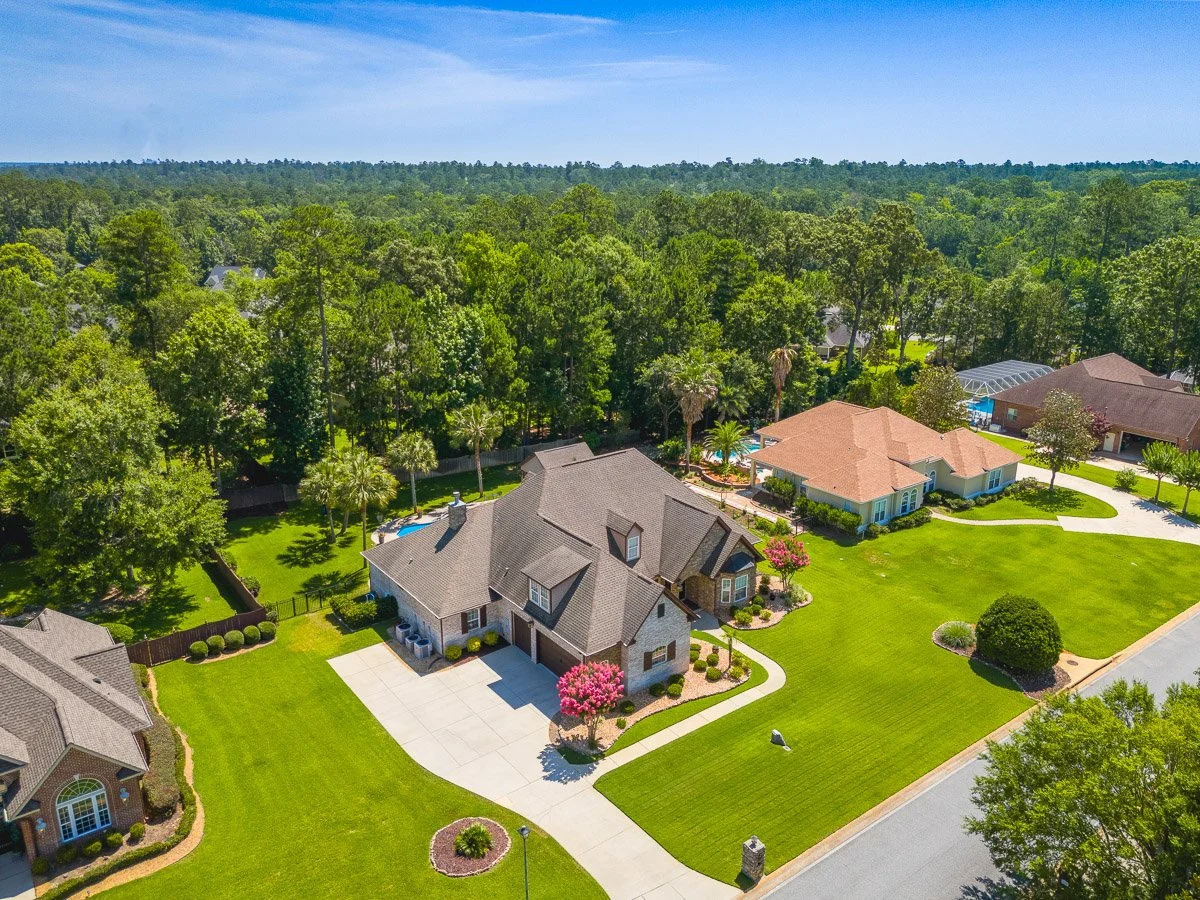 Aerial view of a suburban neighborhood with large houses, green lawns, trees, and a clear blue sky.