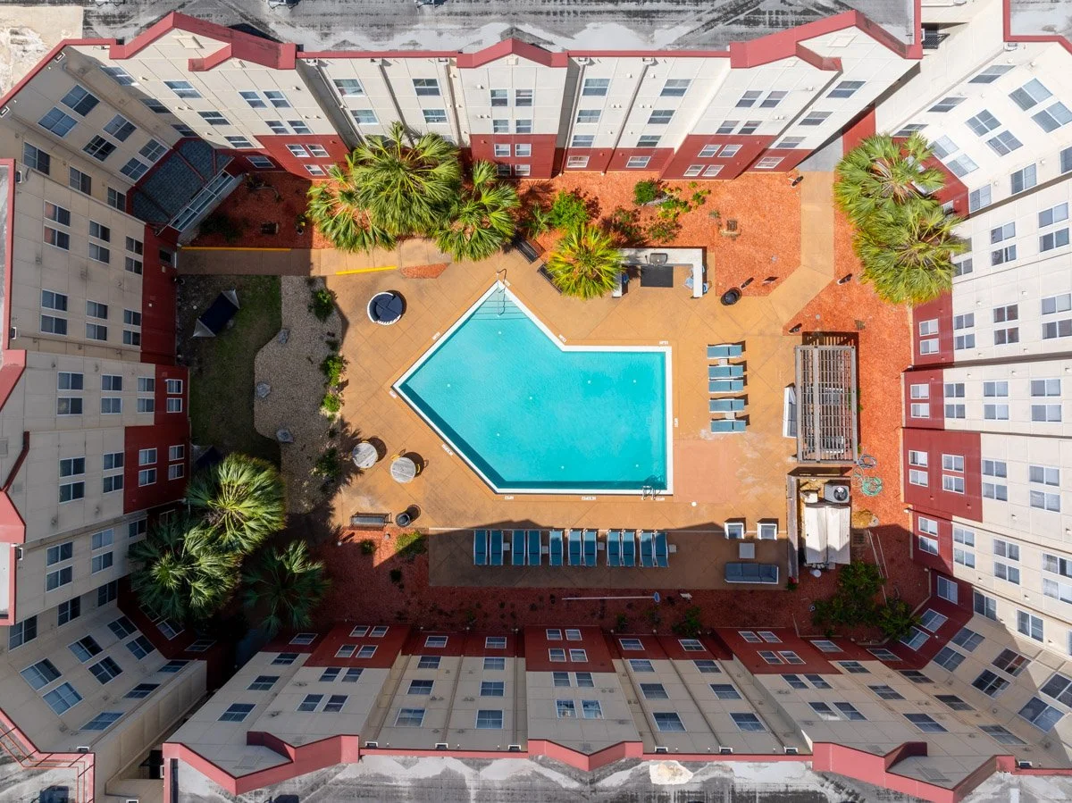 Aerial commercial Real Estate photo of a courtyard swimming pool surrounded by a multi-story residential building with a red and beige exterior, palm trees, lounge chairs, and outdoor furniture.